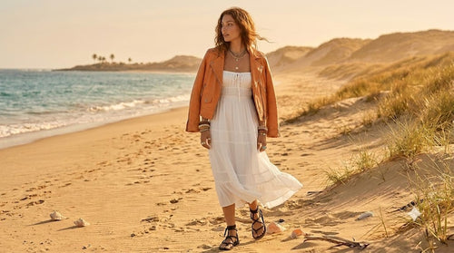 Woman wearing fall boho outfit, a rust leather jacket, white boho maxi dress and bohemian style layered jewelry walking on the beach.