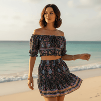 Woman wearing a boho navy blue floral off-shoulder crop top and matching mini skirt with puff sleeves and colorful border detailing on the beach.