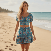 Woman wearing a boho turquoise floral crop top and high-waisted mini skirt matching set with puff sleeves, smocked bodice, and decorative border print on the beach.