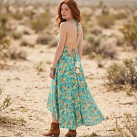 Back of woman wearing a backless teal pastel floral halter maxi dress with a plunging V-neckline, gathered waist, and flowing high-low hem on the beach.