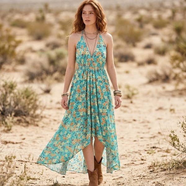 Woman wearing a backless teal pastel floral halter maxi dress with a plunging V-neckline, gathered waist, and flowing high-low hem on the beach.