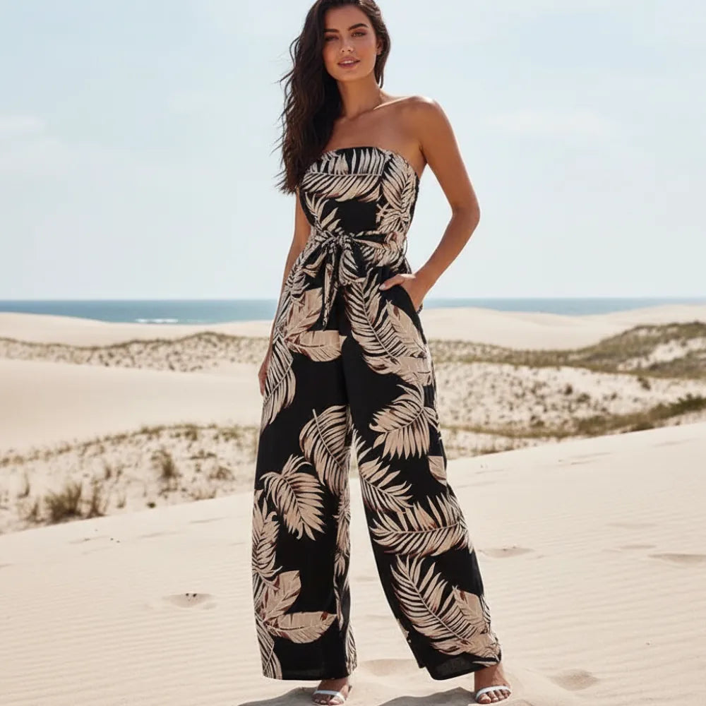 Woman wearing a black boho floral print sleeveless wide leg jumpsuit with a strapless bandeau neckline, tropical palm leaf print, tie waist, and side pockets on the beach.