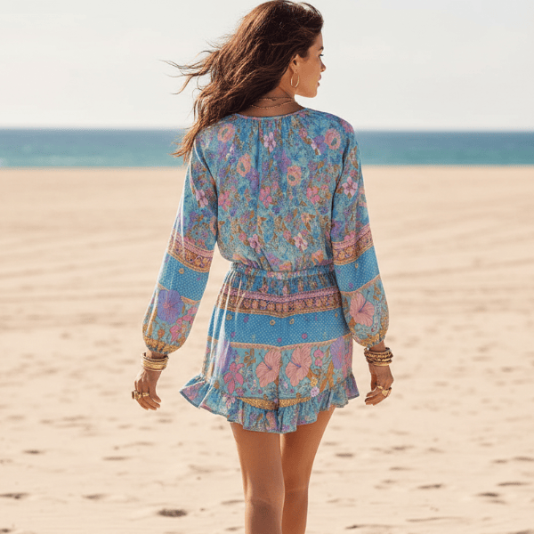 Back of a woman wearing a blue floral boho romper with long balloon sleeves, V-neck button front, tassel drawstring waist, and ruffled shorts for effortless summer style.