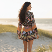 Back of a woman wearing a black floral boho romper with long balloon sleeves, V-neck button front, tassel drawstring waist, colorful floral print, and ruffled shorts.