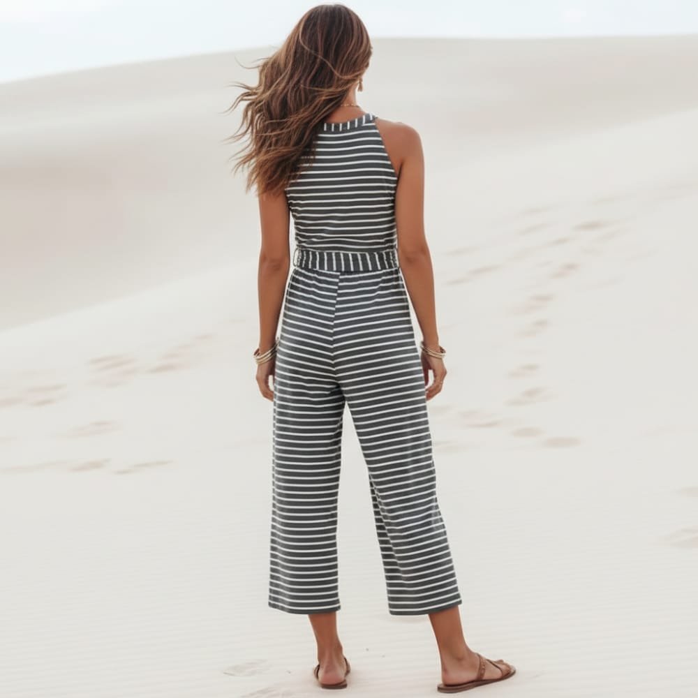 Back of a woman wearing a black boho striped sleeveless wide leg jumpsuit with a round neckline, adjustable tie waist, cropped pants, and relaxed fit on a beach.