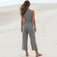 Back of a woman wearing a black boho striped sleeveless wide leg jumpsuit with a round neckline, adjustable tie waist, cropped pants, and relaxed fit on a beach.