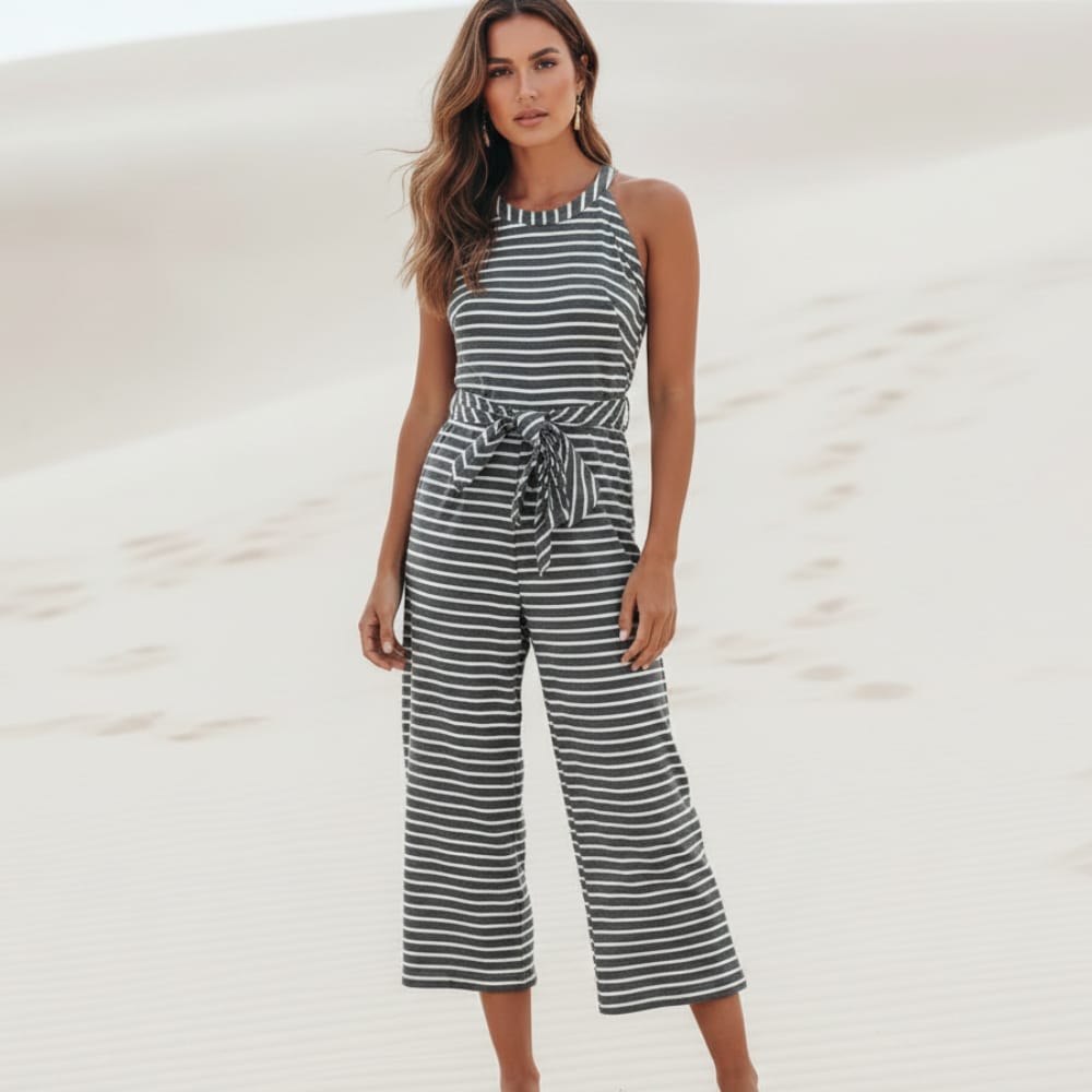 Woman wearing a black boho striped sleeveless wide leg jumpsuit with a round neckline, adjustable tie waist, cropped pants, and relaxed fit on a beach.