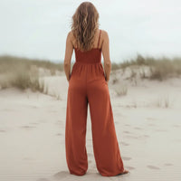 Back of a woman wearing a rust red boho chic wide leg jumpsuit with a smocked bodice, spaghetti straps, side pockets, and flowing full-length pants on a sandy beach.