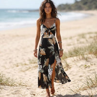 Woman wearing a black floral cutout maxi dress with spaghetti straps, front tie bow, V-neckline, and side slit on the beach.