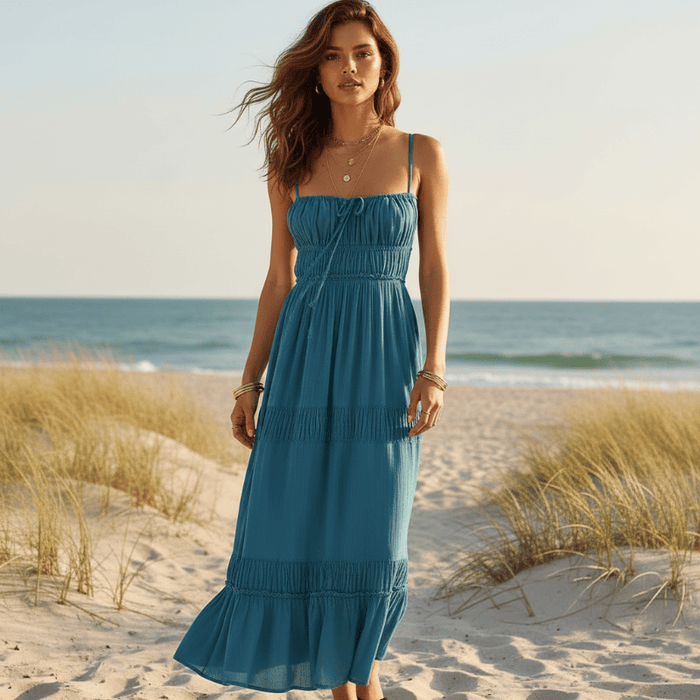 Woman wearing a teal blue boho backless tie-back maxi dress with an open back, flowing A-line silhouette, elastic waist, and ankle-length hem on the beach.