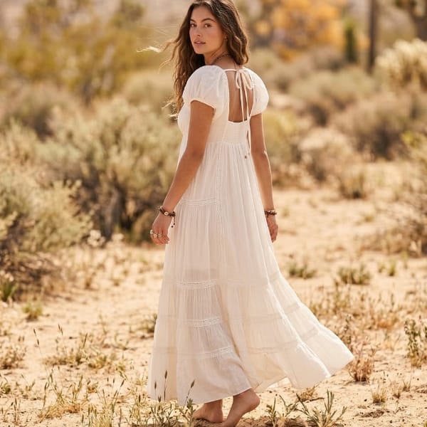 Back view of an off-white cotton maxi dress featuring an open-back cutout, tie closure, puff sleeves, and flowing tiered skirt.