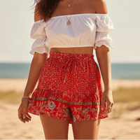 Woman wearing Scarlet Blossom boho floral shorts with a high waist drawstring waistband, lightweight rayon fabric, relaxed flowy fit, feminine ruffle hem, and a vintage-inspired floral border in vibrant scarlet red.