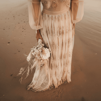 Bride showing the bottom of a boho Champagne lace beach wedding dress with embroidered tulle, illusion long sleeves, deep V neckline, low back, flowing A-line skirt, and sweep train on the beach.