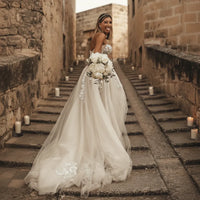 Bride walking upstairs wearing a boho lace wedding dress featuring crystal lace appliqués, sweetheart neckline, off-the-shoulder sleeves, flowing A-line skirt, lace-up corset back, and elegant court train for a romantic outdoor wedding.