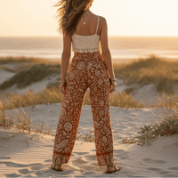 Back view of a woman wearing rust paisley boho wide leg pants with functional side pockets, a high waist drawstring waistband, and a flowy palazzo silhouette paired with a white tank top.