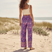 Back of woman wearing Wild Orchid boho wide leg pants with functional side pockets, a high waist drawstring waistband, lightweight rayon fabric, and a flowy purple floral palazzo silhouette with a vibrant botanical print.