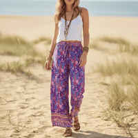 Woman wearing Wild Orchid boho wide leg pants with functional side pockets, a high waist drawstring waistband, lightweight rayon fabric, and a flowy purple floral palazzo silhouette with a vibrant botanical print.