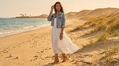 A woman on a sandy beach wearing a long white boho maxi dress with a light blue denim jacket and brown ankle boots.